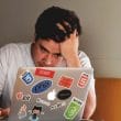 hombre sentado tocándose la cabeza con la mano mientras mira su computadora gris con stickers de colores; hombre con estrés por trabajo