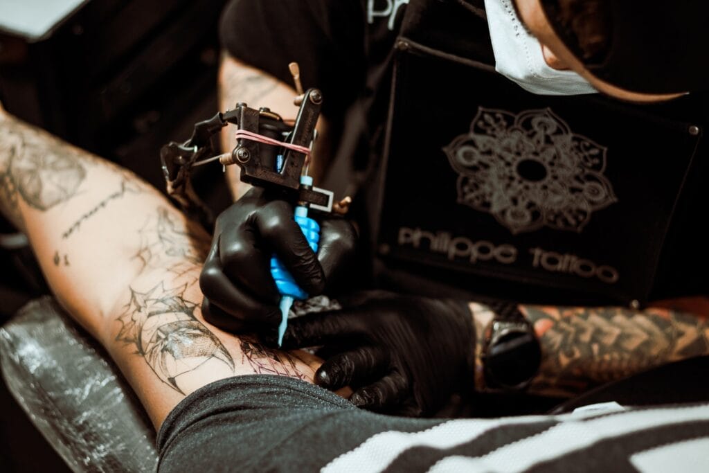 Hombre con camisa, gorra y guantes color negro. En la mano tiene una maquina de tatuar color negra con azul. El hombre se encuentra tatuando un brazo de una persona.