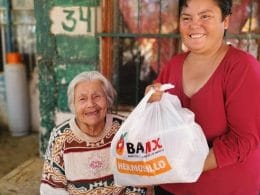 mujer de blusade manga larga roja sostiene bolsa de plástico con alimentos en el interior y a un costado una señora anciana sonríe