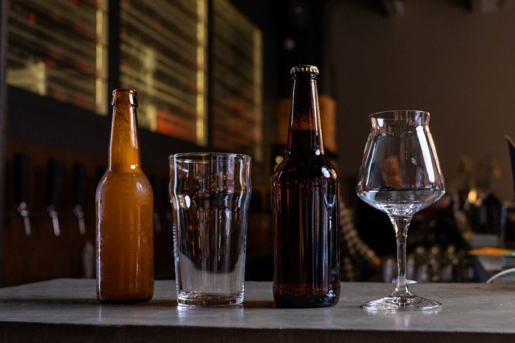 De lado derecho botella de cerveza color ambar, despues un vaso de cristal, enseguida otra botella de cerveza y al final una copa de vidrio.