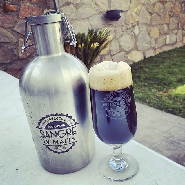 En la parte izquierda una botella grande de acero inoxidable con logotipo de letras color negro, en el lado derecho una copa grande con cerveza oscura.