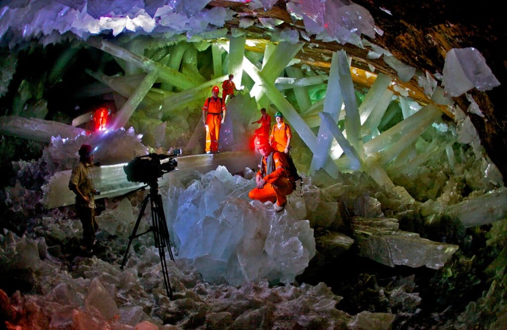 Cueva llena de cristales color blanco con 7 personas vestidos de color naranja, en el centro hay una cámara con tripie color negro.