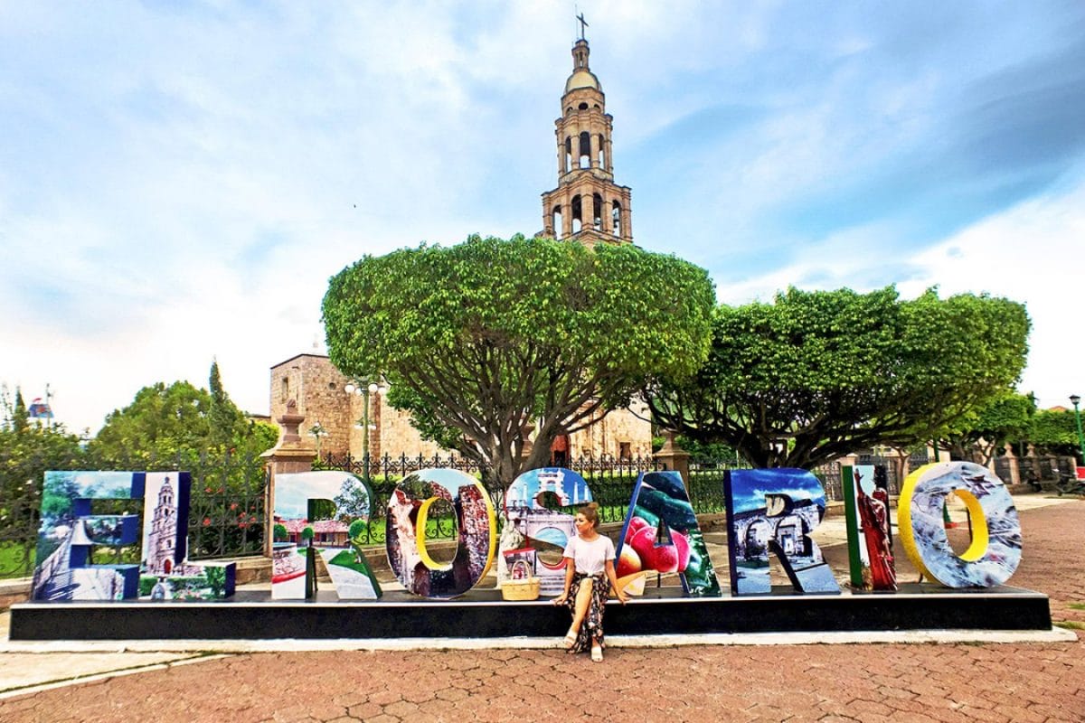 Plaza con letras que dicen "El Rosario" de diferentes colores e imagenes. En esta letras gigante se encuentra una chica con falda color negra y blusa blanca.