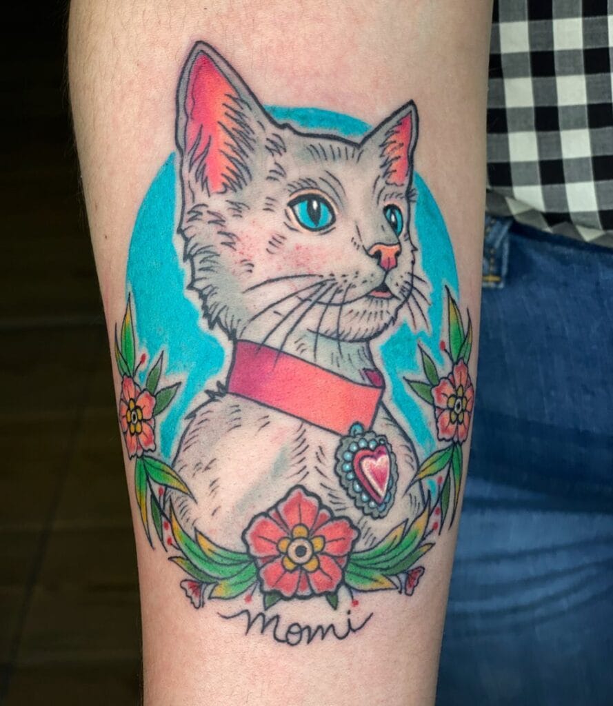 Tatuaje de gato color blanco-gris con un collar rojo y un corazón de dije. En la parte de abajo del gato se encuentran flores color rojo-naranjas y con letras color negro "momi".