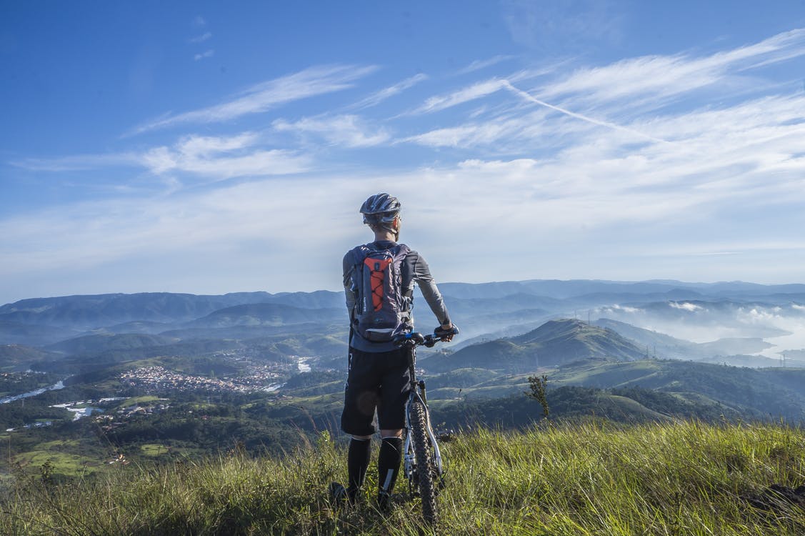 Imagen ilustrativa de un ciclista de espaldas, mientras sostiene su bicicleta y mira hacia las montañas.